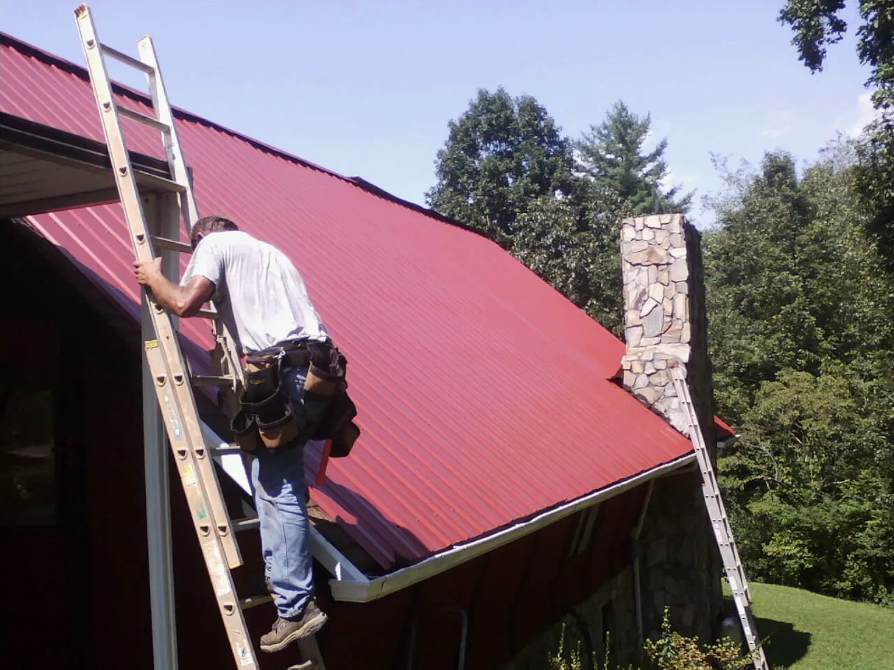 Roofer on a ladder beside a red metal roof and stone chimney — Soffit & Fascia Repair by Roofing Brothers in Orange